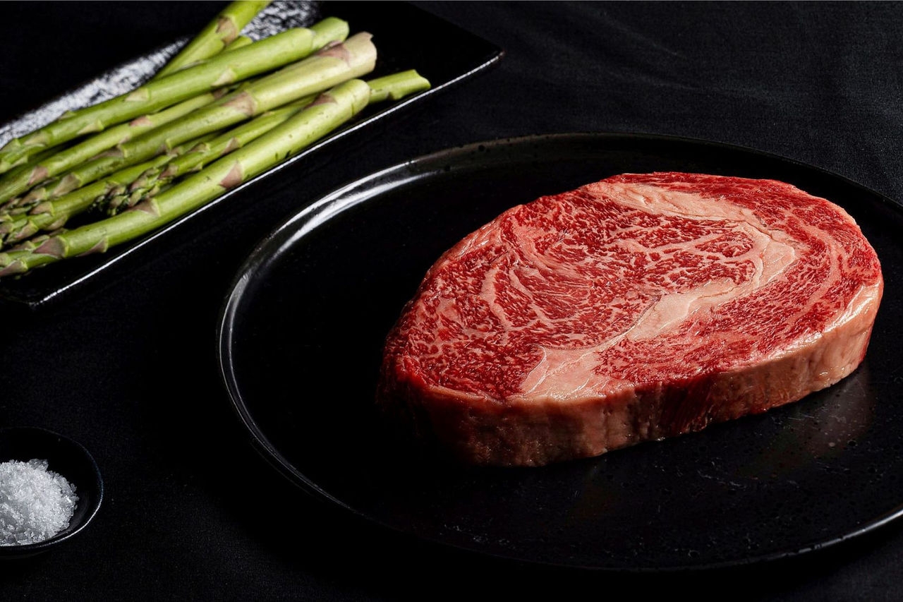 A raw beef steak and asparagus spears are displayed on black plates with a small black bowl of salt
