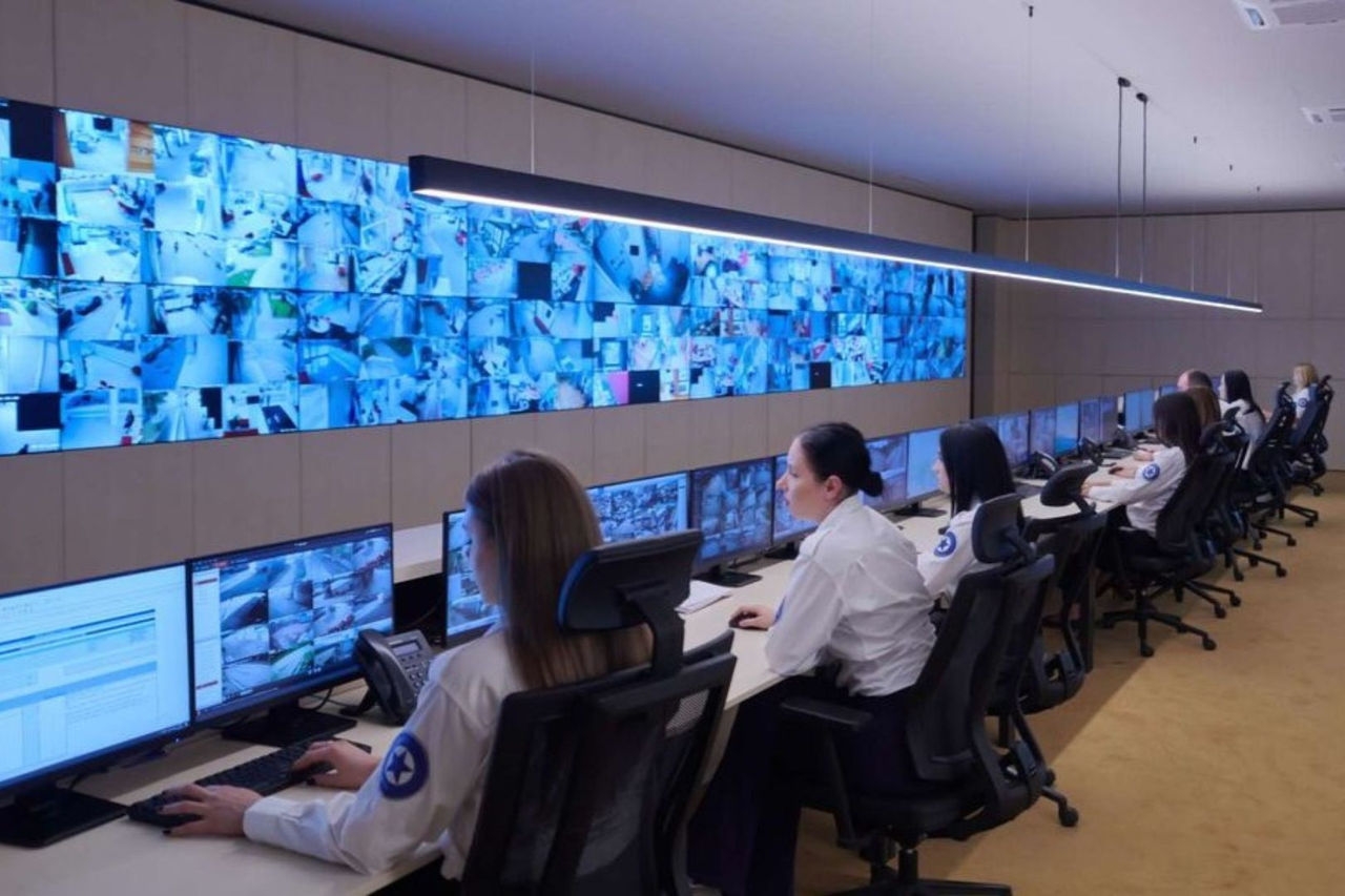 a number of security personnel sit facing a wall of computer displays while typing on computer keyboards