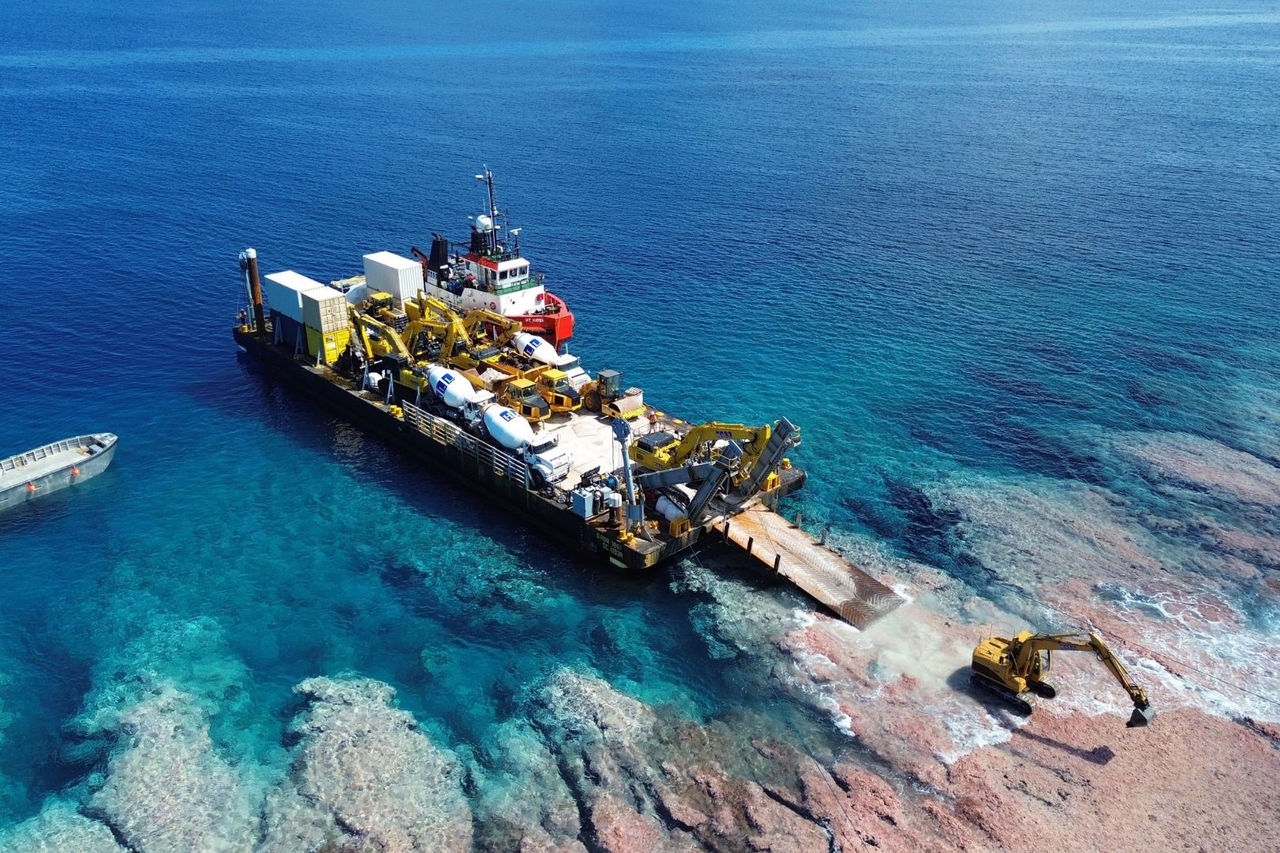 a sea-going barge carrying a range of large construction machinery lands ashore with an excavator driving across rocks