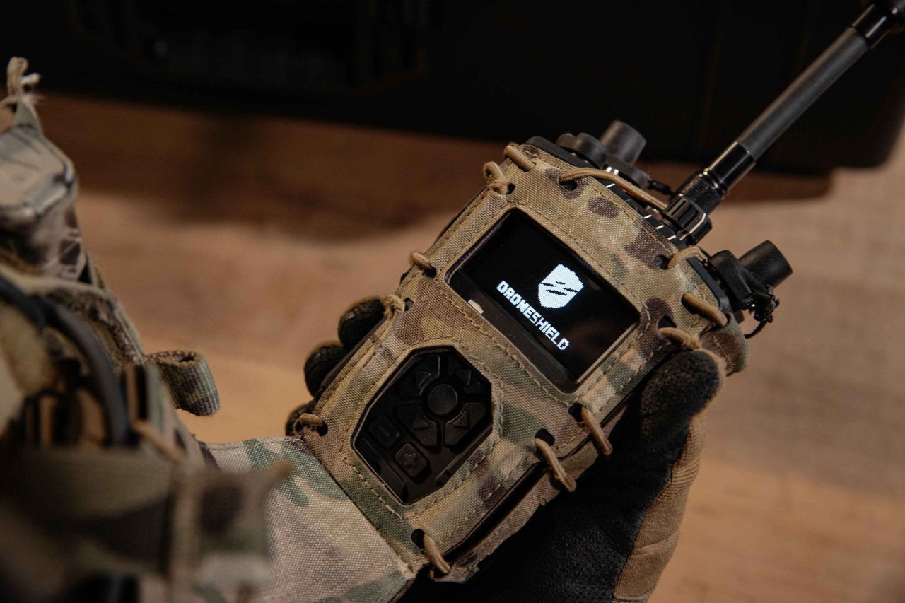 A handheld terminal with knobs and antennae held by someone in a miltary camo clothing