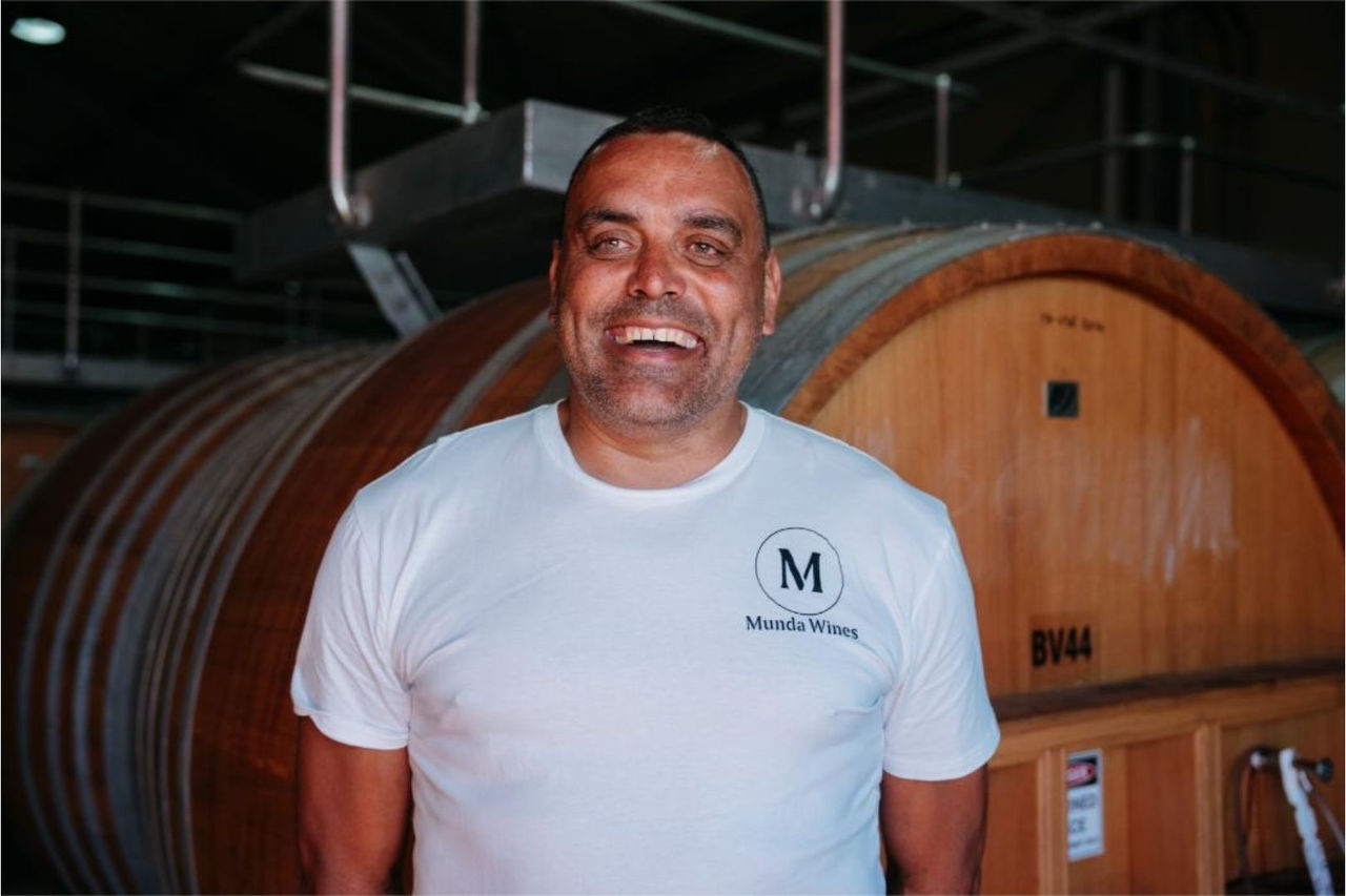 a smiling man wearing a white t-shirt stands in front of a large wine barrel
