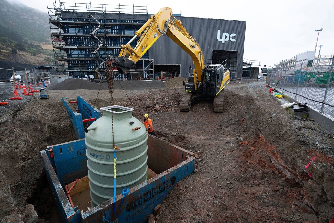 a yellow backhoe arm lowers a green wastewater container into the ground with a metal building under construction in the background