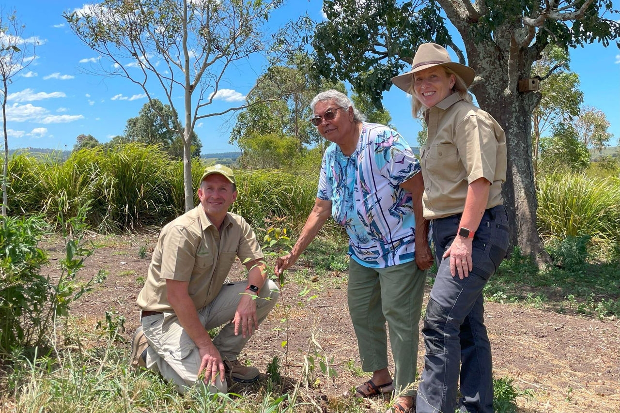 a male crouching and two women standing surrounded by bushland and trees with a walking trail behind them