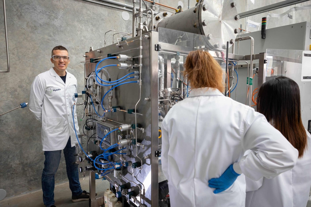 Three lab technicians stands next to a stainless steel frame with pipe and wires connected to it