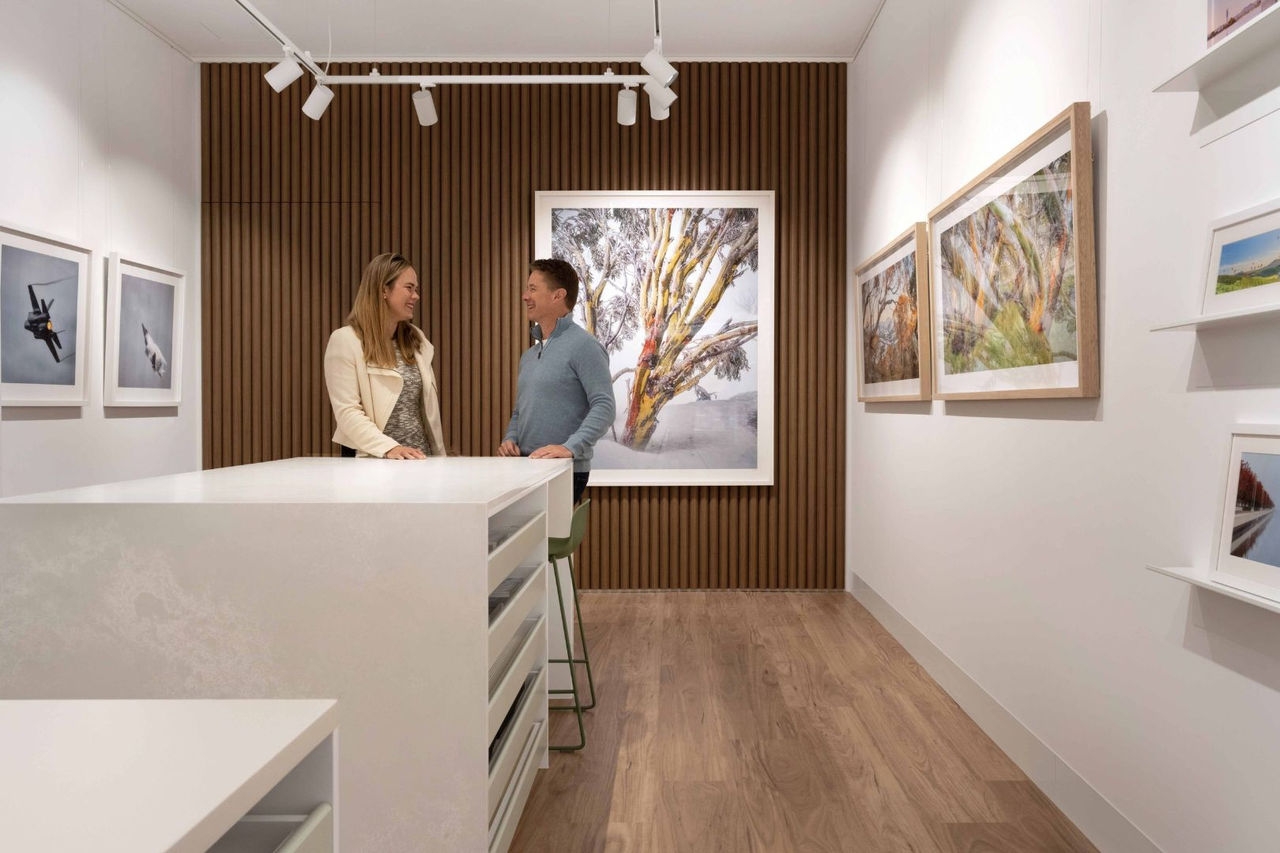 A male and a female stand in a gallery with framed pictures hanging on the walls