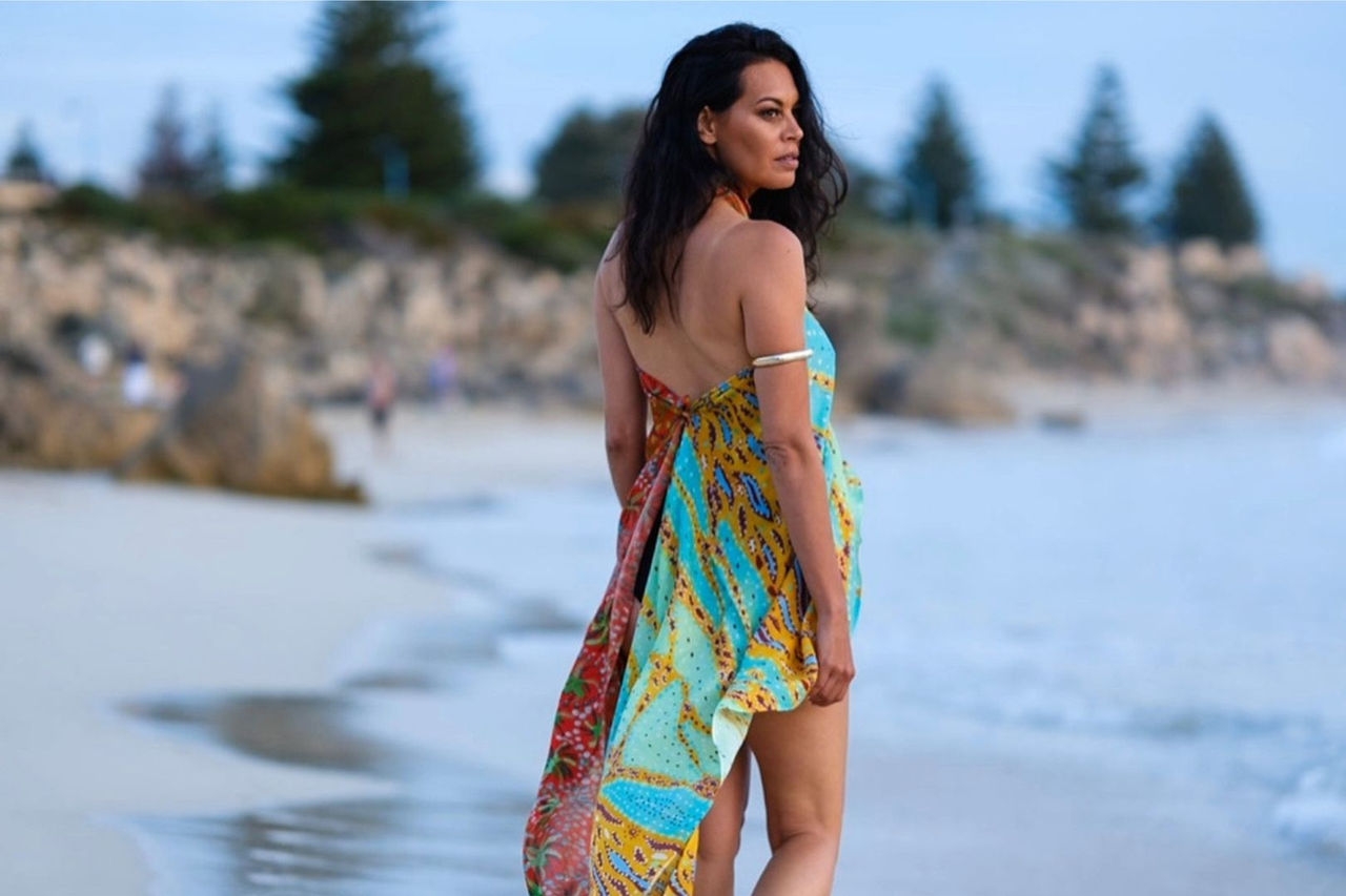 a woman models a dress on a beach with water, dunes and trees in the background