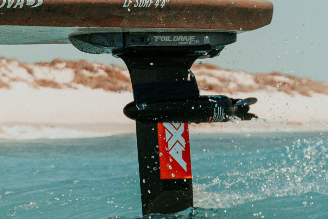 An electric propulsion motor under a board, glides just above the water at a beach with dunes in the background