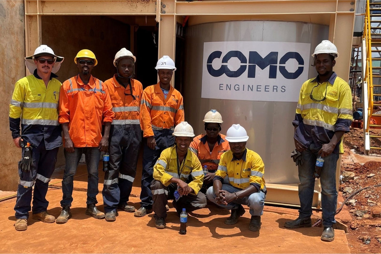 A group of men wearing high visibility work clothes pose in front of steel framework and a metal tank