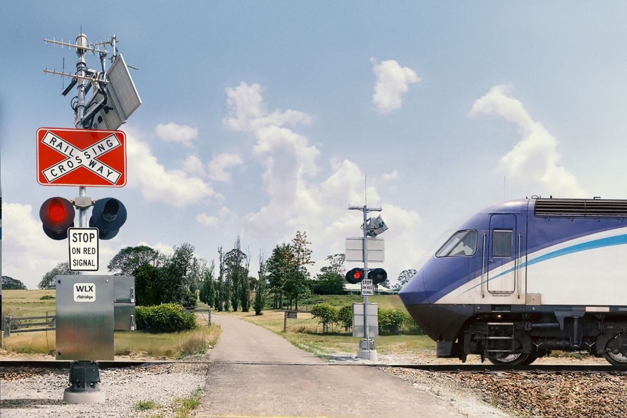 the front of a blue and white train engine rolls across a level road crossing passing a warning sign and flashing lights