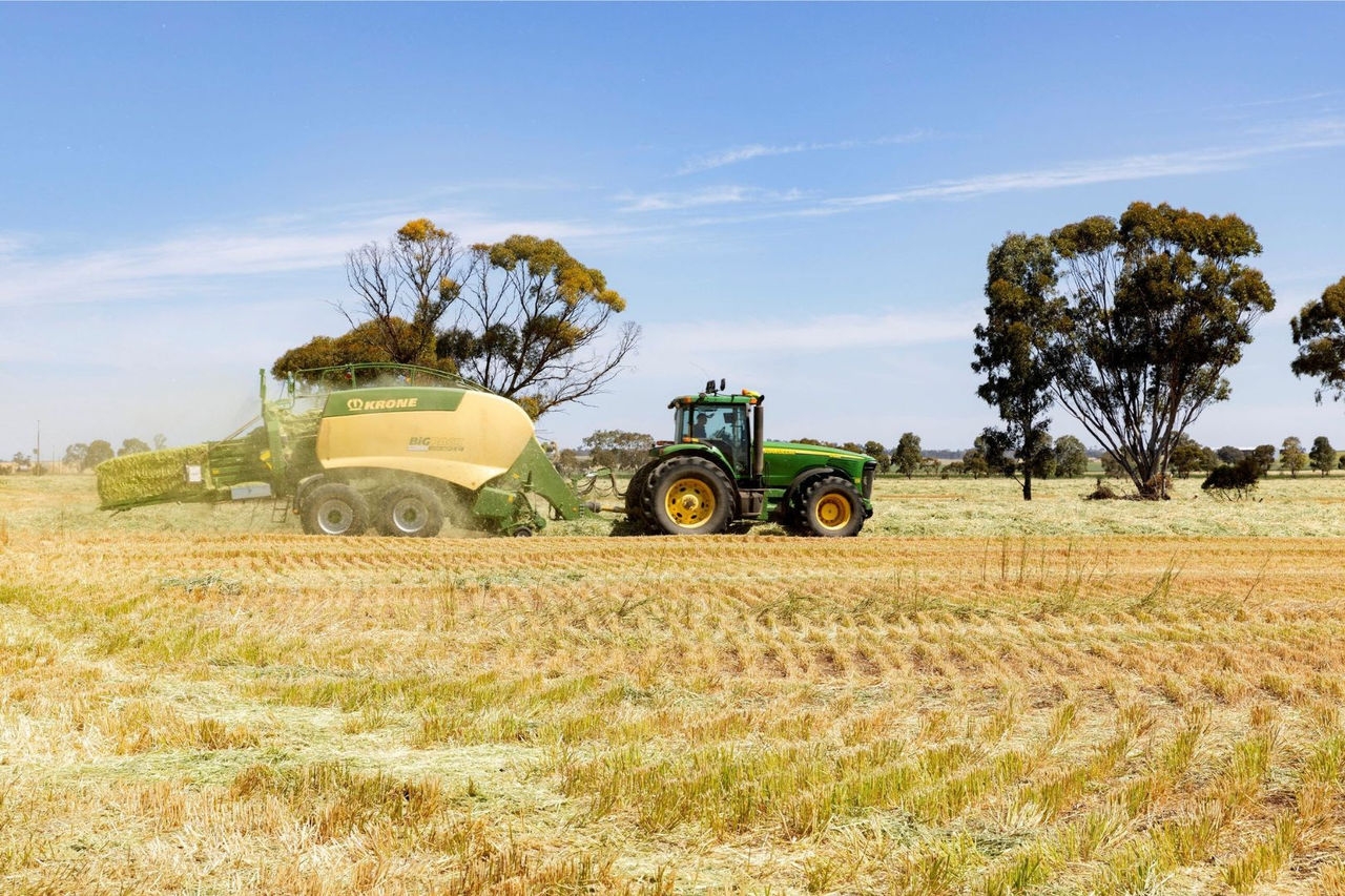 a tractor tows a harvester machine through a field with random trees and blue sky in the background
