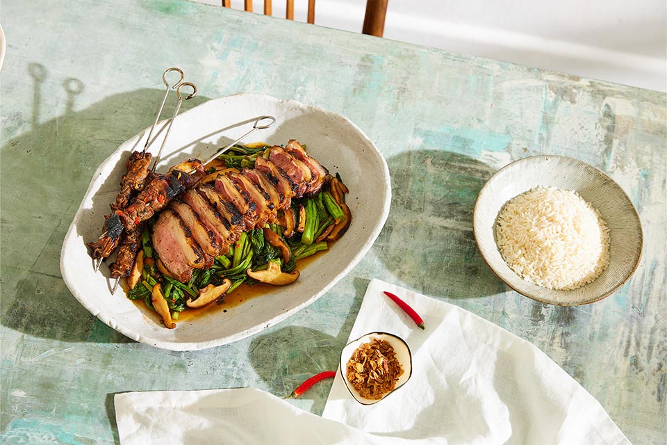 Cuts of BBQ lamb on a serving platter of greens with rice, chilli and dried shallots