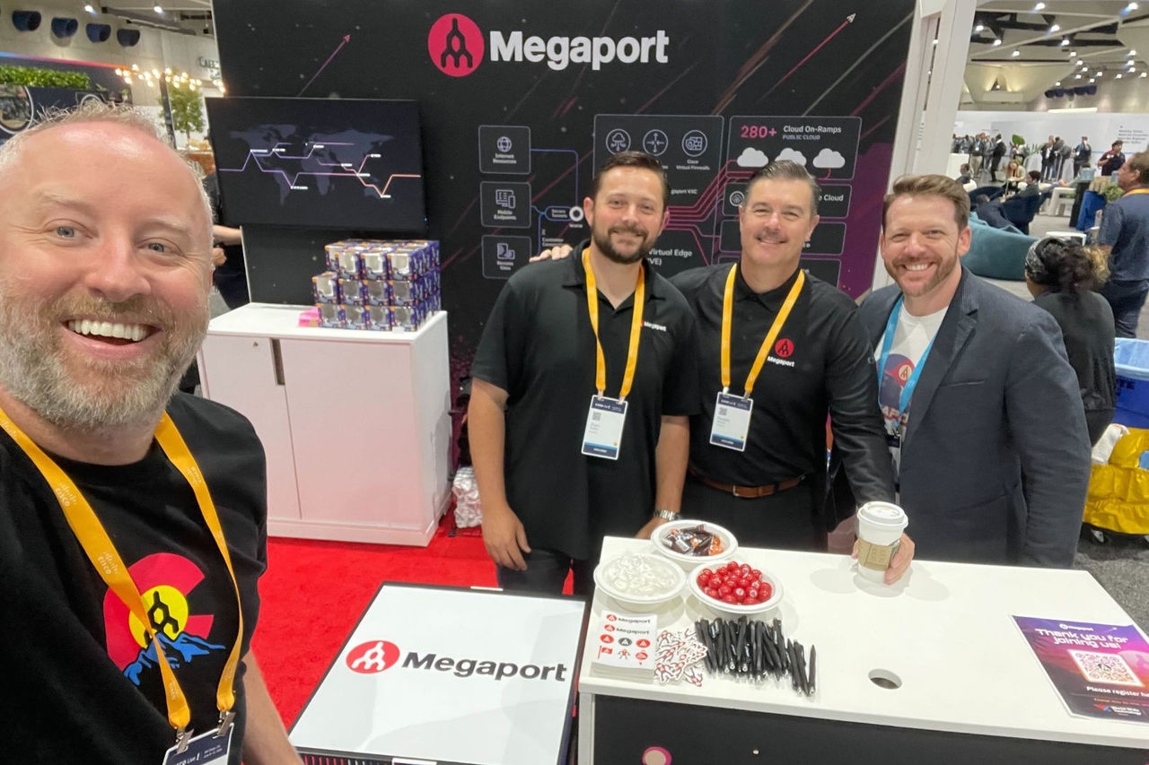 Four men pose for the camera while manning a booth at a technology tradeshow