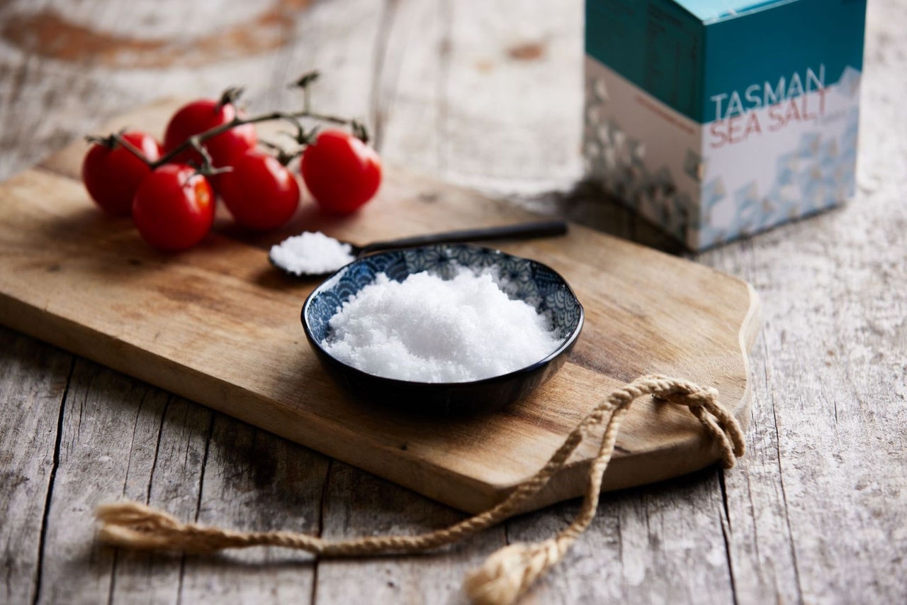 A wooden serving board with a bowl of white salt with truss tomatoes behind.