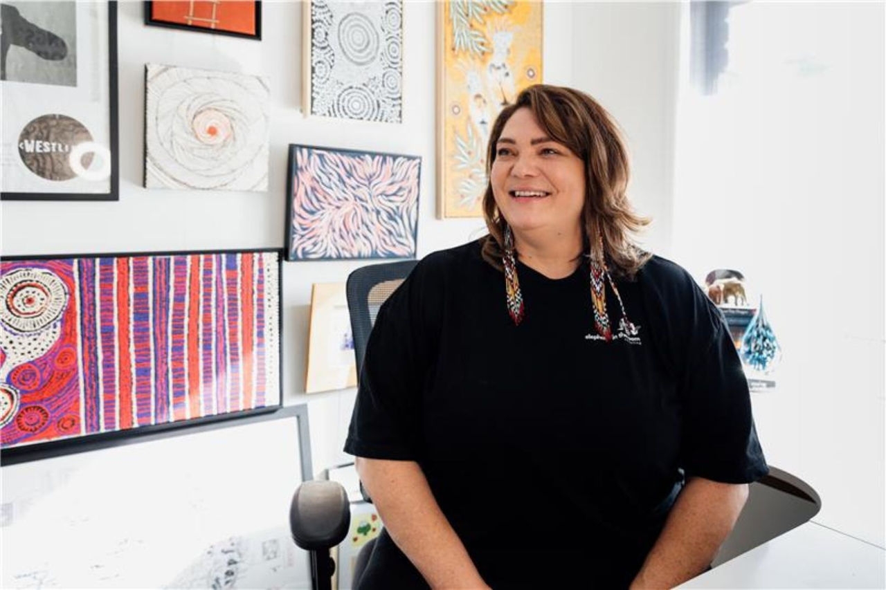 a woman wearing a black top sits in an office chair with First Nations artwork hanging on a white wall